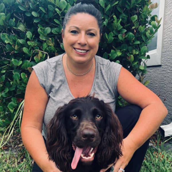A smiling woman with dark hair pulled back, wearing a gray shirt, sits outside in front of green bushes, hugging a happy brown dog with long ears and its tongue out—showcasing the special bond between pet and veterinarian.